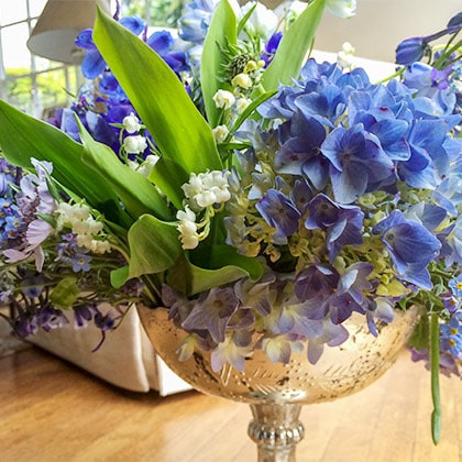 A mercury glass compote filled with Lily of the Valley, blue Belladonna delphinium and blue hydrangeas.