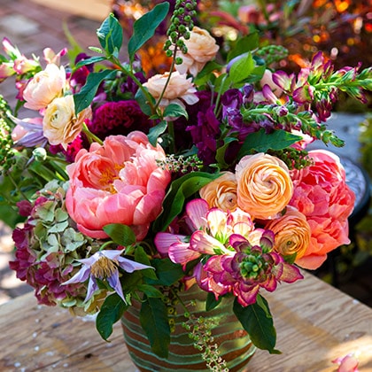 A large flower arrangement of burgundy dahlias, ranunculus, foxglove, peonies, lavender clematis and hydrangeas in a green and gold glass vase.